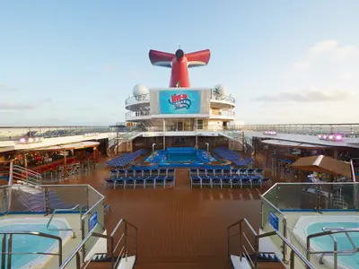 Weitwinkel des leeren Pooldecks der Carnival Sunrise am Tag. Im Zentrum Pool und Liegestühle. Ein großer Bildschirm mit "Dive-In Movies" und roter Schornstein im Hintergrund. Bars und Jacuzzis seitlich.