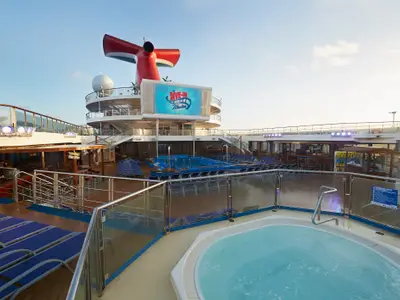 Ansicht des Pooldecks der Carnival Sunrise am Tag. Im Vordergrund ein sprudelnder Whirlpool, im Hintergrund der Hauptpool mit zahlreichen blauen Liegen. Ein großer Bildschirm zeigt 'Dive-In Movies'. Der rote Schornstein ist sichtbar.