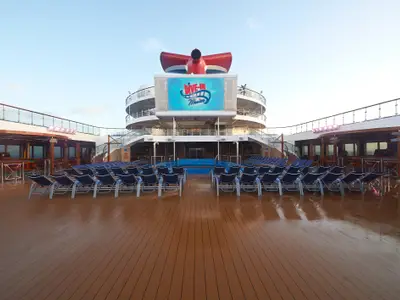 Weitwinkelansicht des Pooldecks der Carnival Sunrise bei Tageslicht. Blaue Liegestühle, Pool und große Leinwand ('Dive-In Movies') sind zu sehen. Bars und Decks im Hintergrund. Der nasse Holzboden reflektiert.