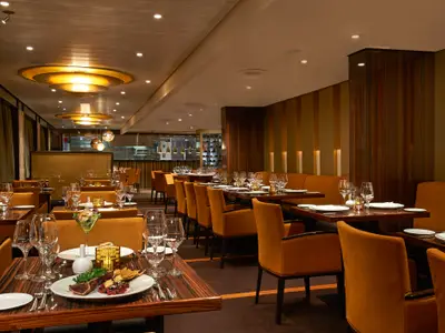 Ein elegantes Restaurant mit Reihen von goldbraunen Samtstühlen und dunklen Holztischen, gedeckt mit Geschirr und Gläsern. Warme Beleuchtung von der Decke und den Wänden erzeugt eine luxuriöse Atmosphäre. Eine offene Küche ist im Hintergrund sichtbar.