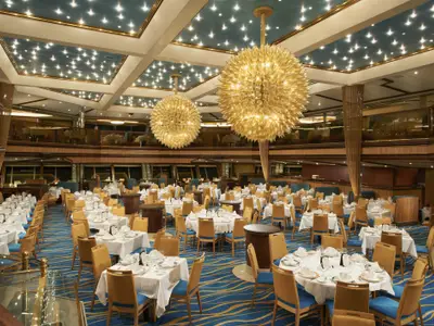 Ansicht eines mehrstufigen Restaurants an Bord der Carnival Sunrise mit zahlreichen festlich gedeckten Tischen und eleganten Stühlen. Zwei große, goldene Kronleuchter hängen von der Decke, die mit vielen kleinen Lichtern als Sternenhimmel gestaltet ist. Einladende und luxuriöse Atmosphäre.
