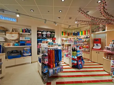 Ein heller Laden auf der Carnival Sunrise mit der Aufschrift 'Carnival Adventures', Verkaufsregalen voller Kleidung, Taschen, Lanyards und einer bunten Süßigkeitenbar mit rot-weiß gestreiftem Boden.