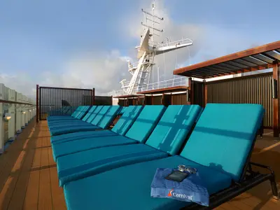 Blick über mehrere Reihen leerer, blauer Liegestühle auf einem Holzdeck der Carnival Sunrise. Im Hintergrund ist die weiße Maststruktur und blauer Himmel mit Wolken zu sehen. Eine Sonnenbrille und ein Buch liegen auf einem Handtuch auf dem vordersten Stuhl.