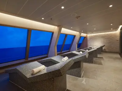 Ein moderner Ruheraum auf der Carnival Sunrise mit mehreren gekachelten, beheizten Liegen, jede mit einem gerollten weißen Handtuch. Große Fenster bieten einen Blick auf das tiefblaue Meer bei Dämmerung oder Nacht. Die Decke ist mit eingelassenen Lichtern versehen, und eine Wand ist mit strukturiertem Stein verkleidet.