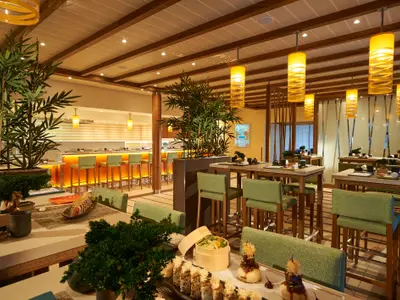 Asiatisches Spezialitätenrestaurant auf der Carnival Sunrise. Helle Tische, grüne Hochstühle, eine beleuchtete Sushi-Bar und große Bambuspflanzen prägen den Raum. Warme gelbe Hängelampen. Ohne Personen.