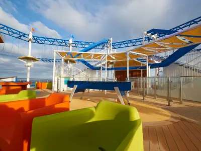 Ein bunter Außenbereich auf einem Deck der Carnival Sunrise mit einem blauen Seilgarten, einem Foosballtisch und leuchtend grünen und orangefarbenen Sitzmöbeln. Blauer Himmel mit Wolken im Hintergrund.