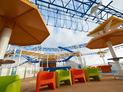 Abenteuerpark auf dem Oberdeck der Carnival Sunrise. Blauer Seilparcours, gelbe Sonnensegel. Bunte Stühle in Grün und Orange auf dem Teakholz-Deck unter orangen Sonnenschirmen. Leichter bewölkter Himmel.