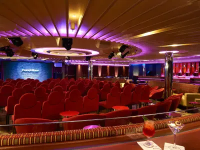Innenansicht eines modernen Comedy-Clubs an Bord der Carnival Sunrise. Rote Sessel reihen sich vor einer Bühne mit dem "Punchliner Comedy Club"-Logo und lila Backsteinwand. Rechts eine Bar mit Spirituosen und lila Akzentbeleuchtung. Zwei Cocktails im Vordergrund.