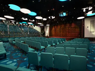 Blick in einen modernen, leeren Theatersaal auf der Carnival Sunrise. Die Bühne mit rotem Vorhang ist rechts. Zahlreiche Reihen tealfarbener Sessel stehen auf einem gemusterten Teppich. Darüber erheben sich weitere Ränge mit Bestuhlung. Die Decke ist dunkel mit vielen Strahlern und großen, runden, leuchtenden Paneelen in Weiß und Hellblau.