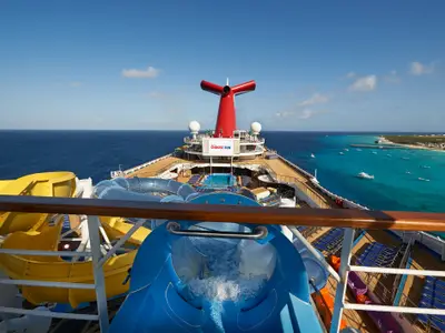 Blick vom Oberdeck der Carnival Sunrise mit gelben und blauen Wasserrutschen, Pool und dem roten Schornstein. Im Hintergrund weites Meer und eine Küstenbucht mit vielen Booten.