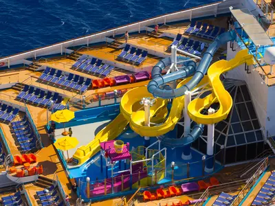 Vogelperspektive auf ein Sonnendeck der Carnival Sunrise mit gelben und blauen Wasserrutschen, Pools und vielen Liegestühlen. Einzelne Passagiere sind sichtbar. Das blaue Meer umgibt sie.