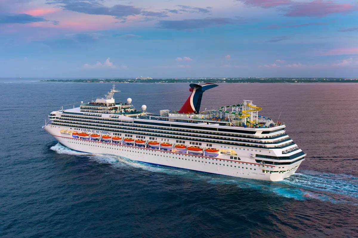 Panorama-Ansicht der weißen Carnival Sunshine auf blauem Meer bei Sonnenuntergang. Rote und blaue Schornsteine, bunte Wasserrutschen, und eine ferne Küstenlinie sind sichtbar. Das Schiff hinterlässt eine Schaumspur.