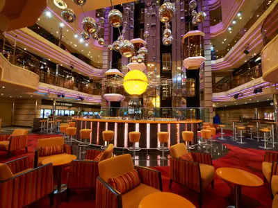 Modernes Atrium der Carnival Sunshine mit einer zentralen Bar unter einer großen, goldenen Sonnen-Lichtskulptur. Orangefarbene Sessel und Tische auf rotem Teppich. Mehrere obere Decks sind sichtbar.