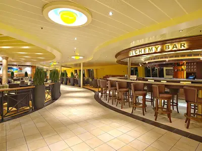 Moderne Alchemy Bar auf der Carnival Sunshine. Langer Bartresen mit braunen Barhockern. Dahinter Sitzbereiche mit hohen Pflanzen und warmer Beleuchtung.