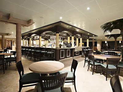 Innenansicht einer stilvollen Bar der Carnival Sunshine mit zentralem Tresen, vielen Sitzgelegenheiten und Palmen. Warme Beleuchtung. Elegante Holz- und Säulendetails.