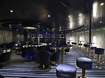 Eine elegante, modern gestaltete Bar und Lounge auf der Carnival Sunshine, dominiert von dunklem Blau und Gold. Der Raum ist mit bequemen Samtsesseln und hohen Barhockern ausgestattet, Tische haben goldene Ränder. Ein gestreifter Boden in Blau- und Schwarztönen zieht sich durch den Raum, ergänzt durch glänzende Lamellenvorhänge und indirekte Beleuchtung.
