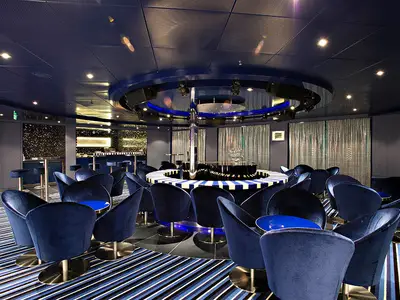 Eine moderne Bar/Lounge auf der Carnival Sunshine, dominiert von dunkelblauen Samtsesseln und runden Tischen. Im Zentrum steht eine auffällige, runde Bühne mit einer polierten Säule. Im Hintergrund sind eine weitere Bar und Vorhänge zu sehen, die eine elegante Atmosphäre schaffen.