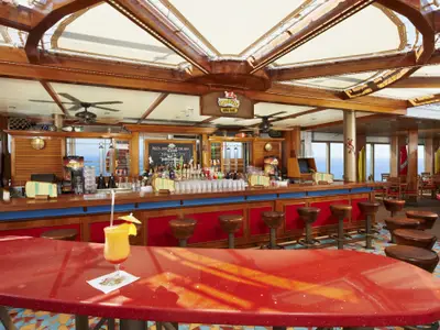 Helle RedFrog Rum Bar auf der Carnival Sunshine mit Holztheke, roten Hockern und großen Fenstern zum Meer. Ein Cocktail steht auf einem roten Tisch im Vordergrund.