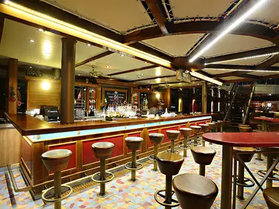 Eine einladende Bar-Lounge auf der Carnival Sunshine mit einer langen Holztheke, zahlreichen Barhockern und roten Tischen. Eine Treppe führt in den oberen Bereich. Stimmungsvolle Beleuchtung schafft eine warme Atmosphäre.