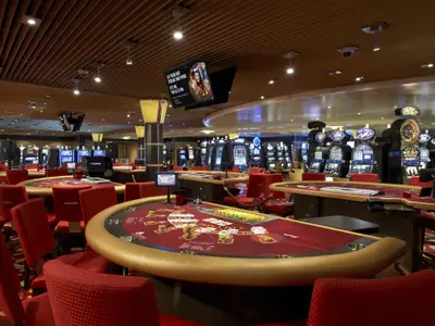 Ein weitlaeufiges Casino auf der Carnival Sunshine mit zahlreichen beleuchteten Spielautomaten im Hintergrund und mehreren roten Spieltischen mit Stühlen im Vordergrund. An der Decke sind Kameras und Beleuchtung angebracht.