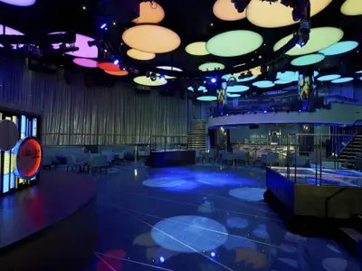 Ein geräumiger Nachtclub auf der Carnival Sunshine, beleuchtet von blauen und farbigen Lichtern. Eine große Tanzfläche, gemütliche Sitzbereiche und eine Bar sind zu sehen, sowie eine Galerieebene.
