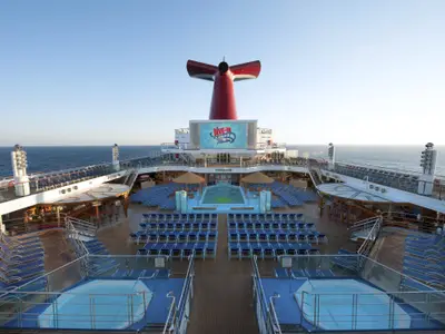 Ein weitwinkeliger Blick von oben zeigt das Pooldeck der Carnival Sunshine mit einem leeren Pool, zahlreichen blauen Liegestühlen und einem großen Bildschirm, der 'DIVE-IN Movies' anzeigt. Im Hintergrund sind das Meer und der blaue Himmel sichtbar.