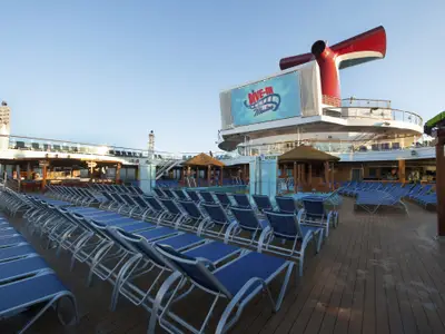 Ein menschenleeres Pooldeck der Carnival Sunshine mit vielen blauen Liegestühlen, einem großen Bildschirm mit 'Dive-In Movies'-Logo, einem roten Schiffsschornstein und mehreren Bars und Pools unter strahlend blauem Himmel.