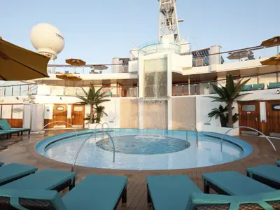 Blick auf den Poolbereich der Carnival Sunshine bei Tageslicht. Ein geschwungener Pool mit Jacuzzi und Wasserfall, umgeben von Holzdeck und blauen Sonnenliegen. Künstliche Palmen und gelbe Sonnenschirme sind sichtbar. Im Hintergrund Kabinen und ein Radom.