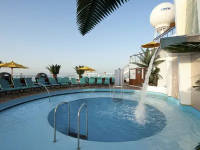 Ein sonniger Poolbereich auf der Carnival Sunshine. Ein runder Pool mit Wasserfontäne und Treppen ist umgeben von türkisfarbenen Liegestühlen, Palmen und gelben Sonnenschirmen. Im Hintergrund ist das Meer sichtbar.
