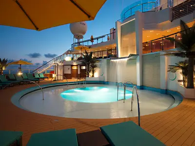 Eine Außenansicht eines beleuchteten Poolbereichs auf der Carnival Sunshine in der blauen Stunde. Ein runder Pool mit Unterwasserbeleuchtung und Treppen ist umgeben von Liegestühlen. Im Hintergrund ein Wasserfall, Palmen und die obere Ebene des Schiffes.