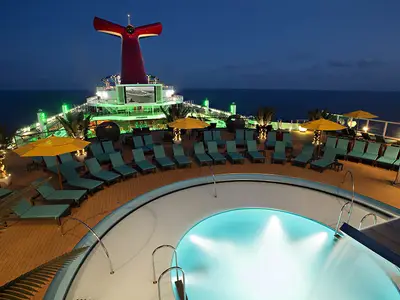 Blick auf das obere Deck der Carnival Sunshine in der Abenddämmerung. Ein beleuchteter Pool mit Sitzgelegenheiten ist im Vordergrund. Dahinter viele Liegestühle, gelbe Sonnenschirme, Palmen und der charakteristische rote Schornstein eines Carnival-Schiffs, beleuchtet von grünen Lichtern. Im Hintergrund ist das dunkle Meer zu sehen.