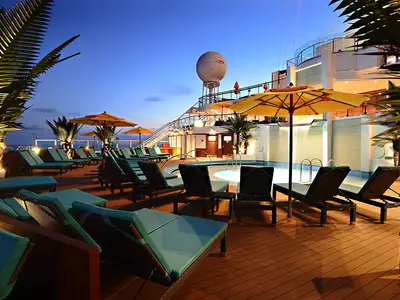 Ein beleuchtetes Sonnendeck der Carnival Sunshine in der Abenddämmerung. Blaue Liegestühle säumen einen hell beleuchteten Pool mit Wasserfall. Im Hintergrund sind Palmen und ein Radom zu sehen.