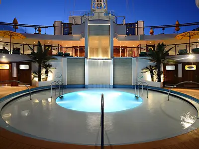 Ein Pooldeck der Carnival Sunshine bei Nacht mit einem leuchtenden runden Pool. Ein beleuchteter Wasserfall stürzt in den Pool. Um den Pool herum sind Palmen und Sitzbereiche auf mehreren Ebenen sichtbar.