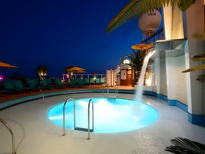 Abendlicher Poolbereich auf der Carnival Sunshine mit leuchtend blauem Pool, Wasserfall und bequemen Liegestühlen. Über dem Pool thronen Sonnenschirme und Palmen, während sich im Hintergrund der Nachthimmel erstreckt.