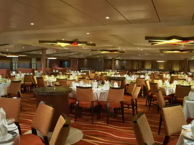 Innenansicht eines eleganten Restaurants auf der Carnival Sunshine. Zahlreiche runde Tische sind mit weißen Tischdecken, Geschirr, Gläsern und Stoffservietten gedeckt. Bequeme, orangefarbene Stühle stehen bereit. Die Decke ist mit dekorativen, farbigen Lichtinstallationen verziert. Der Boden ist mit einem gemusterten, orangefarbenen Teppich ausgelegt. Der Raum ist menschenleer.