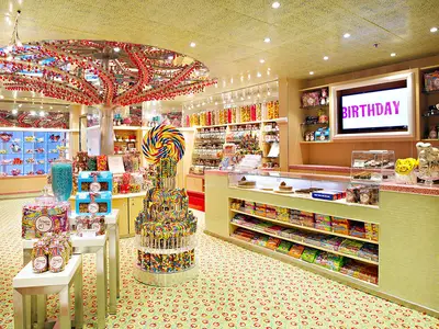 Ein lebhafter Süßwarenladen auf der Carnival Sunshine mit einer bunten Auswahl an Bonbons, Lollipops, Schokolade und Geschenkartikeln. Ein großer Lutscher und ein baumähnliches Dekor mit roten Kugeln dominieren den Raum. Ein Bildschirm zeigt das Wort 'BIRTHDAY' in großen rosa Buchstaben.