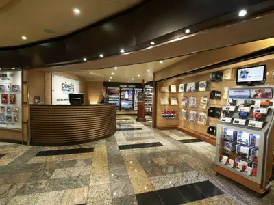 Innenansicht der Pixels Gallery auf der Carnival Sunshine. Ein halbrundes Holzpult, Displays mit Kameras, gerahmte Fotos und Souvenirs sind zu sehen. Der Boden ist mit gemusterten Marmorfliesen ausgelegt.