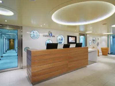 Blick auf die helle und moderne Rezeption des Cloud 9 Spa. Ein langer Holztresen steht im Vordergrund. Dahinter eine weiße Wand mit runden Vitrinen und dem Spa-Logo. Ein Flur führt zu weiteren Behandlungsräumen.
