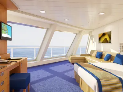 Geräumige Suite der Carnival Sunshine mit blauem Teppich, einem Doppelbett, einem L-förmigen Sofa und großen Fenstern mit Blick auf das offene Meer.