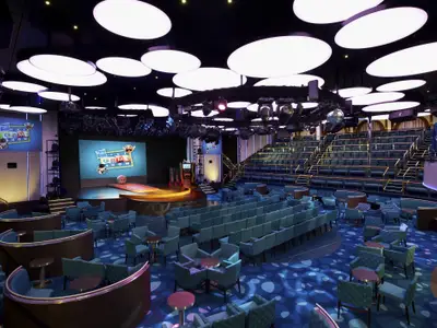 Modernes Theater der Carnival Sunshine für Game Shows. Bühne mit Hasbro-Logo auf Bildschirmen. Türkisfarbene Sitzplätze und Tische. Decke mit runden Lichtern und Scheinwerfern.