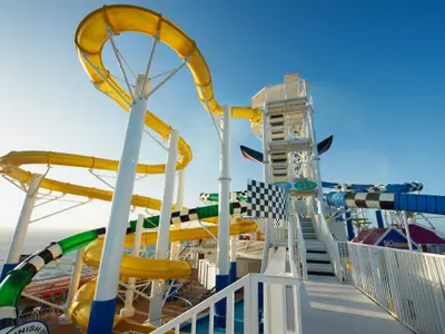 Auf dem Oberdeck der Carnival Sunshine sind bunte Wasserrutschen zu sehen. Gelbe, grüne und blau-weiß gestreifte Röhren schlängeln sich um einen weißen Treppenturm. Blauer Himmel und Meer im Hintergrund.