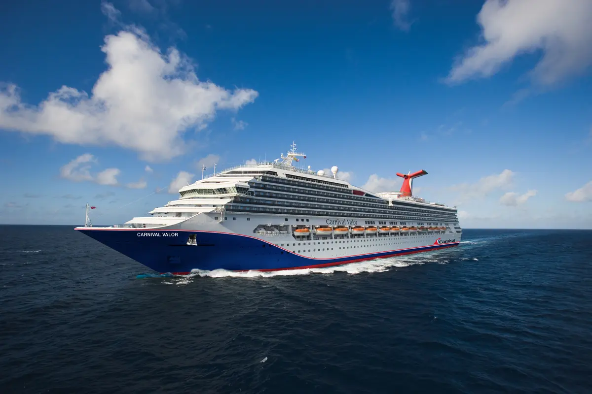 Die Carnival Valor fährt majestätisch auf blauem Meer unter einem strahlend blauen Himmel mit weißen Wolken. Das Schiff ist weiß, blau und rot lackiert, mit mehreren Decks und orangefarbenen Rettungsbooten an der Seite. Eine Bugwelle ist sichtbar.