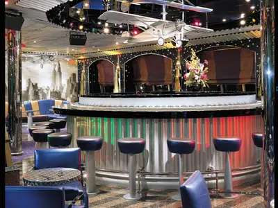 Stilvolle Bar auf der Carnival Valor: Theke im Piano-Design mit blauen Barhockern. New York Skyline als Wandtapete und ein Modellflugzeug an der Decke.