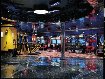 Innenansicht einer lebhaften Spielhalle auf der Carnival Valor mit blauer Beleuchtung. Zahlreiche Arcade-Automaten wie Rennspiele und Tanzautomaten säumen den Raum. Links steht ein gelber Zugwaggon als Dekoration. Der Boden ist glänzend und reflektiert die Lichter.