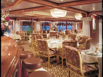 Ein opulentes Restaurant an Bord der Carnival Valor. Zahlreiche, festlich gedeckte Tische mit goldenen Stühlen, üppigen Blumenarrangements und großen Fenstern zum Meer sind zu sehen.