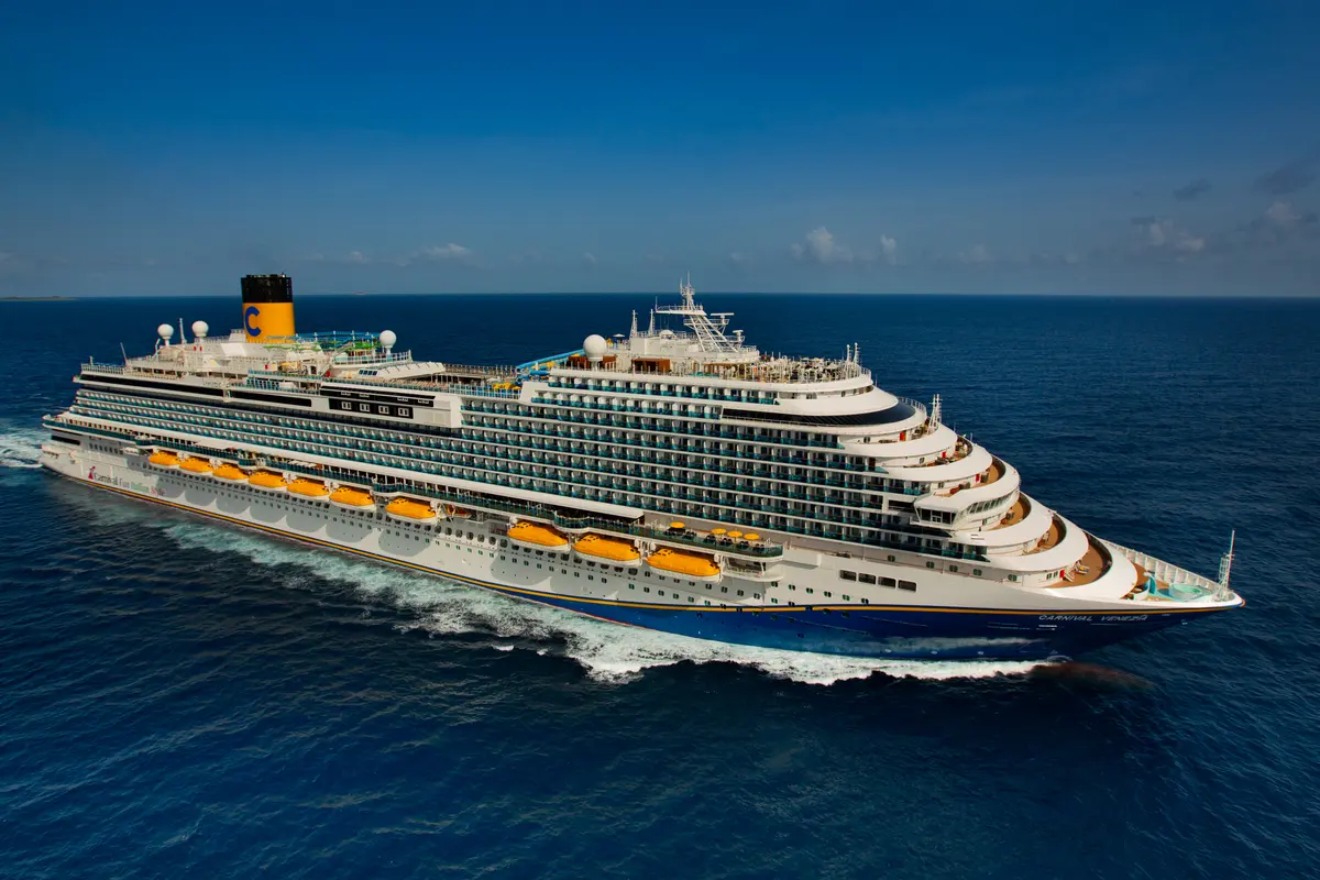 Die große, elegante Carnival Venezia fährt auf dem tiefblauen Meer und hinterlässt eine weiße Gischtspur. Der Himmel ist klar und blau mit wenigen Wolken. Das Schiff ist von oben-hinten fotografiert und zeigt die gelb-schwarzen Schornsteine sowie die blau-weiße Bemalung.