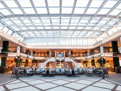Weitwinkelaufnahme eines eleganten Atriums der Carnival Venezia mit großem Glasdach und vielen Sitzgruppen. Mehrere Ebenen mit Restaurants und Bars sind sichtbar. Eine zentrale Treppe mit Brunnen bildet den Mittelpunkt.