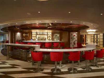 Eine stilvolle Bar auf der Carnival Venezia mit leuchtend roten Barhockern an einer U-förmigen Theke. Hinter der Theke ist eine beleuchtete Bar mit vielen Flaschen zu sehen. Die Decke ist mit dekorativen Holzelementen verziert.