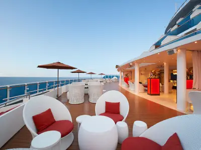 Panoramablick auf das Sonnendeck der Carnival Venezia. Weiße Lounge-Möbel mit roten Kissen stehen unter braunen Sonnenschirmen am Meer. Im Hintergrund eine stilvolle Bar mit Kunst.