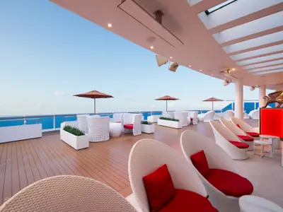 Modernes Außen-Deck auf der Carnival Venezia. Weiße Lounge-Möbel, rote Kissen und Sonnenschirme. Holzdeck, Meer, blauer Himmel. Goldene Skulpturen auf roten, beleuchteten Sockeln.